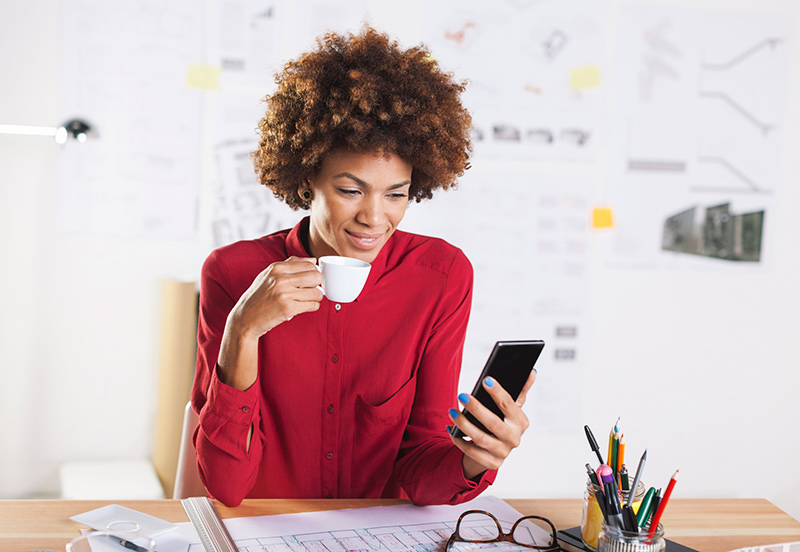 young woman drinking at desk using mobile phone